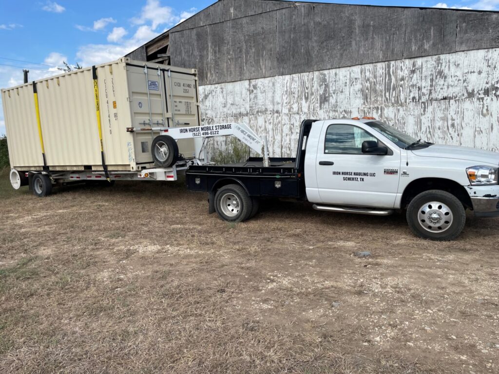 A white flatbed truck is towing a large beige storage container in front of a weathered barn.